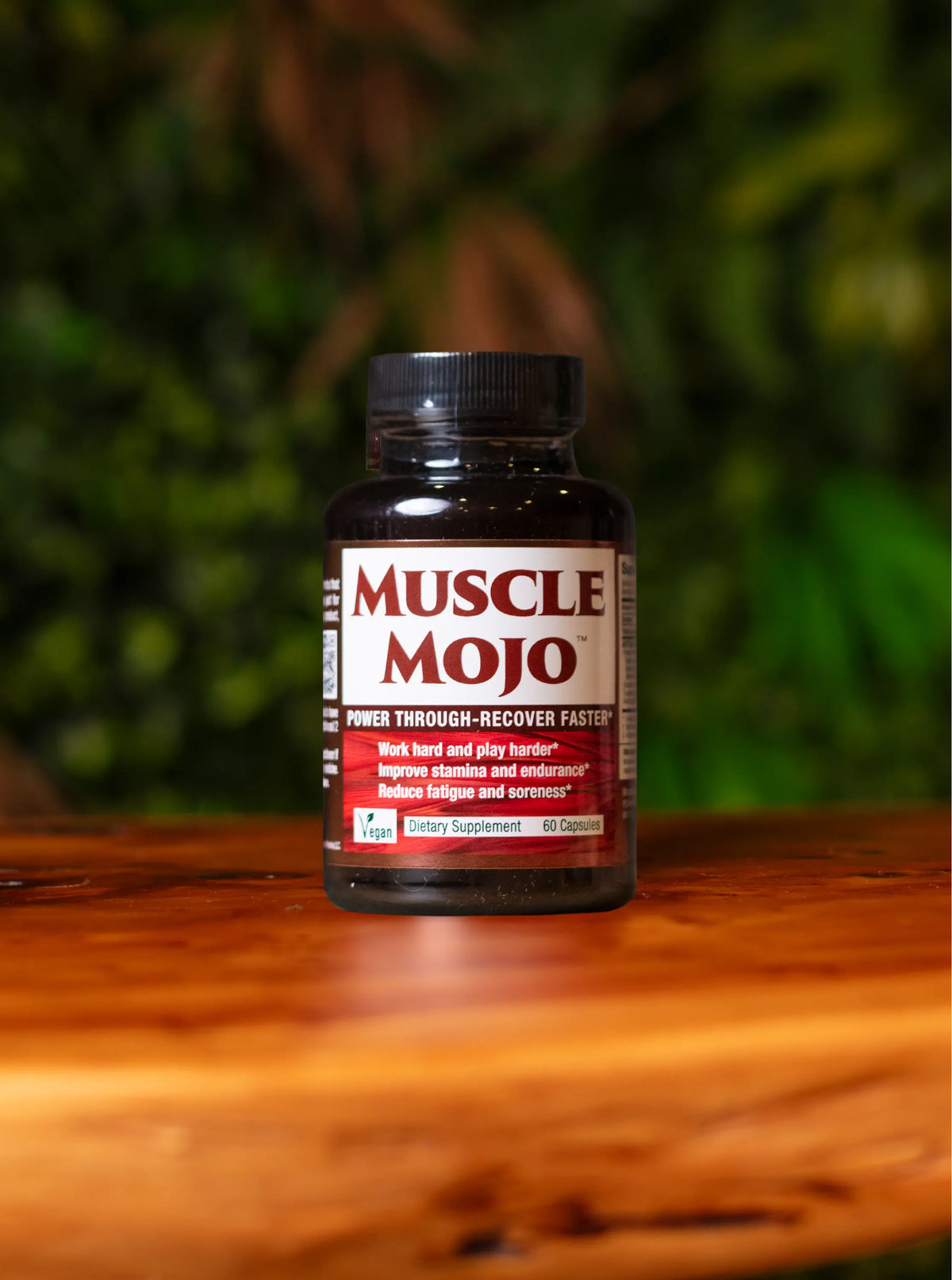 A bottle of Muscle Mojo dietary supplement placed on a wooden surface with a blurred green foliage background.