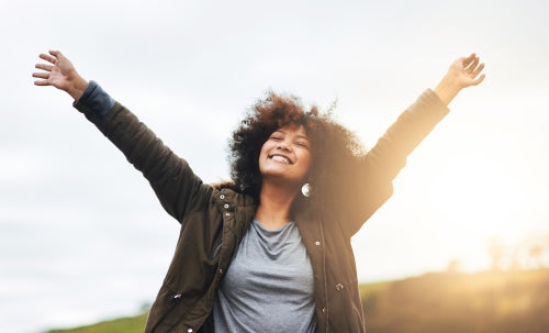 A woman with curly hair stands outdoors with her arms raised in the air, smiling and looking upward, wearing a dark olive green jacket over a gray t-shirt against a bright, sunlit sky.