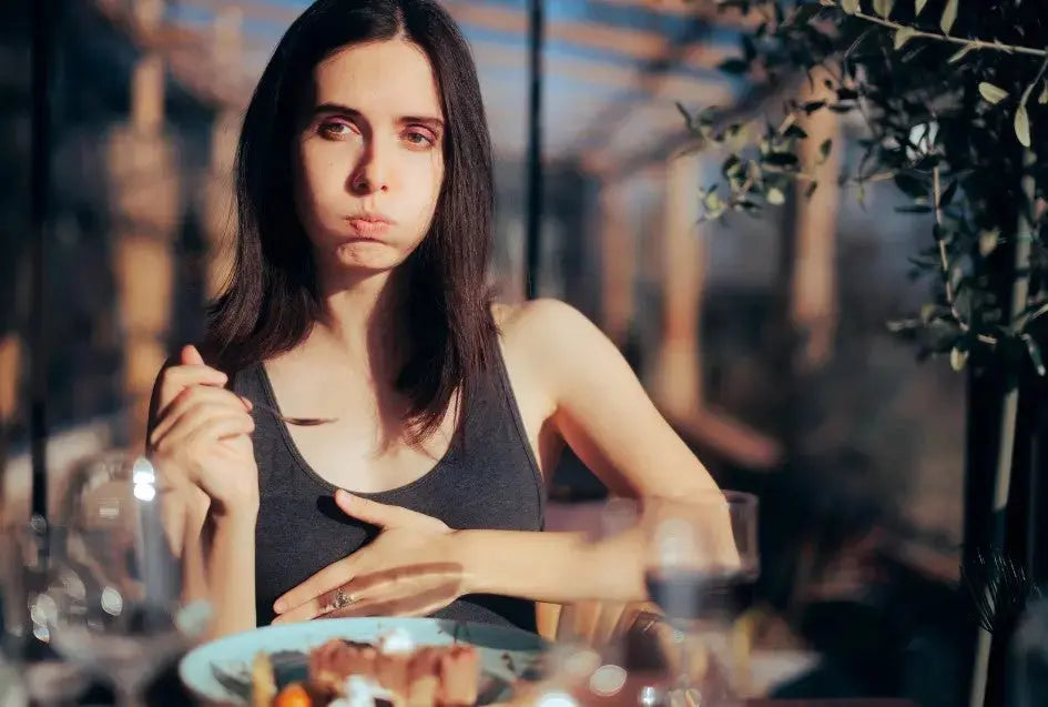 A woman sitting in front of a plate of food holds her hand to her stomach and looks uncomfortable
