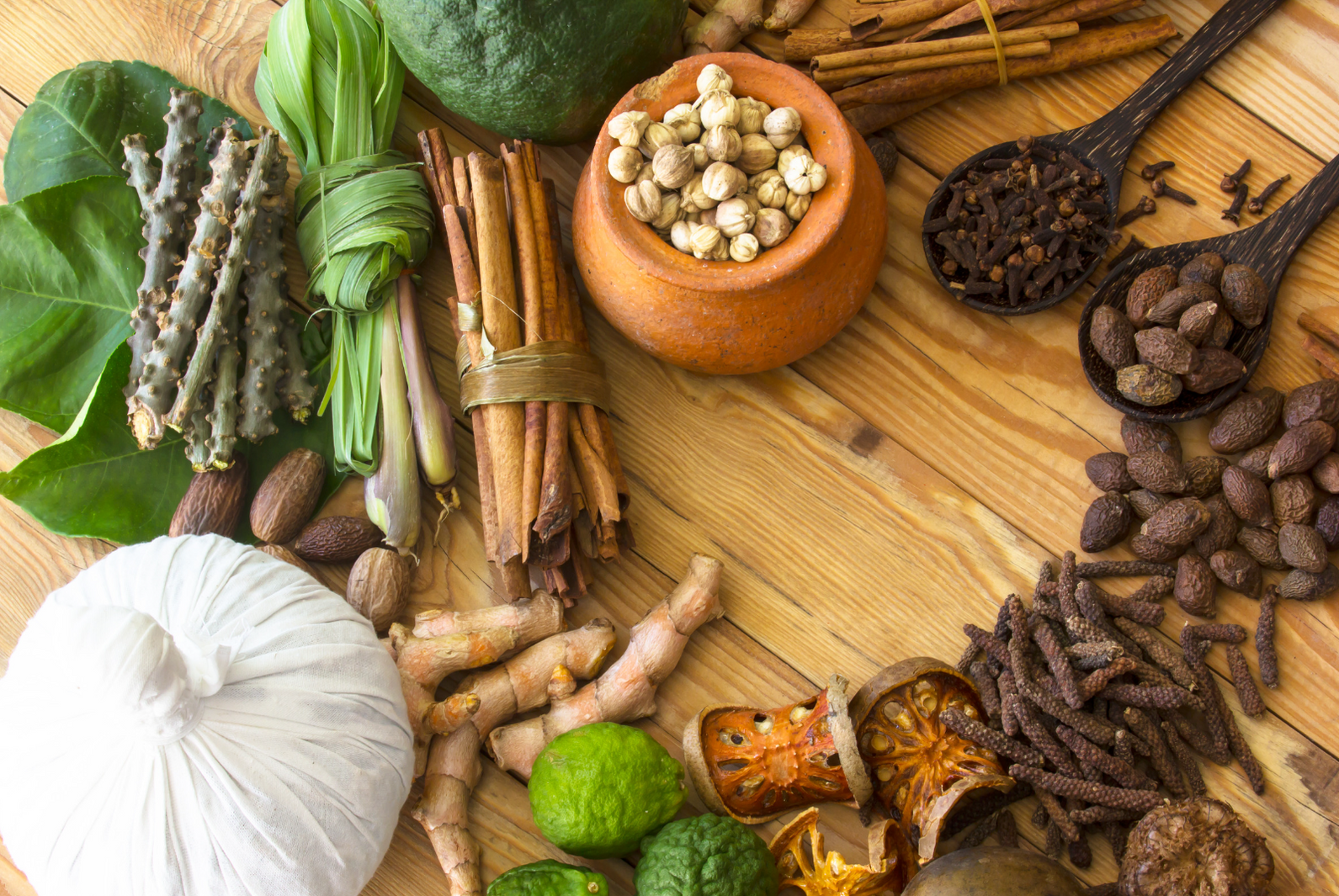 A variety of herbs and spices are laid out on a wooden surface, including long pepper, cloves, cinnamon, fresh turmeric, and more.