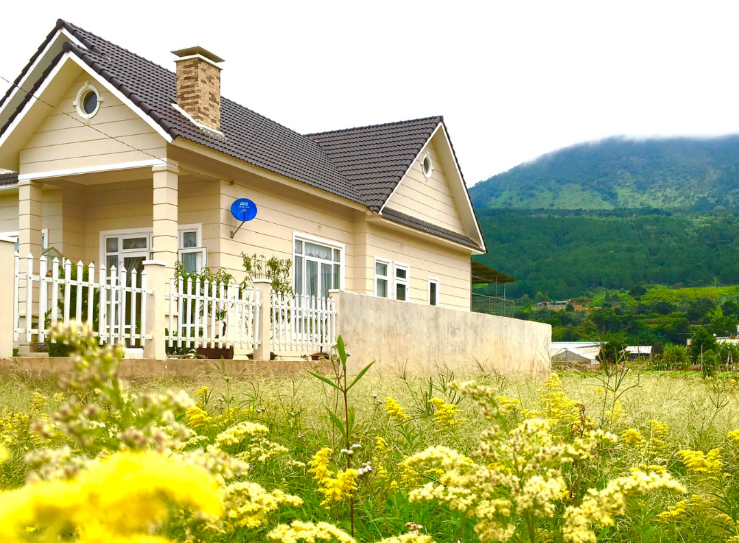 A home with beige clapboard siding and a dark gabled roof sits next to a field of yellow flowers. Mountains and trees are visible in the distance.
