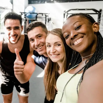 A group of four diverse, smiling people in a gym, with one man giving a thumb's up sign