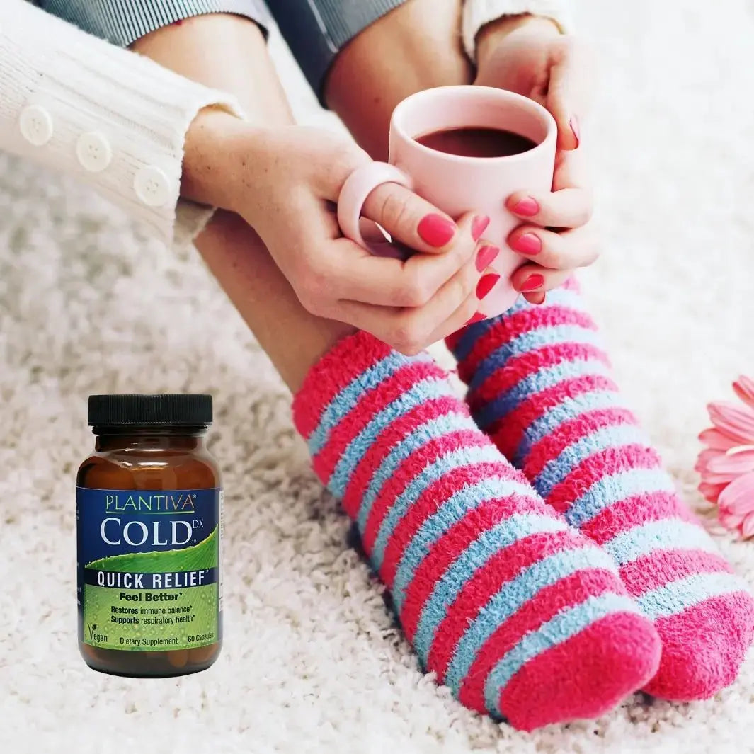 Person wearing colorful socks holding a mug with a bottle of Plantiva Cold Dx on a carpet.