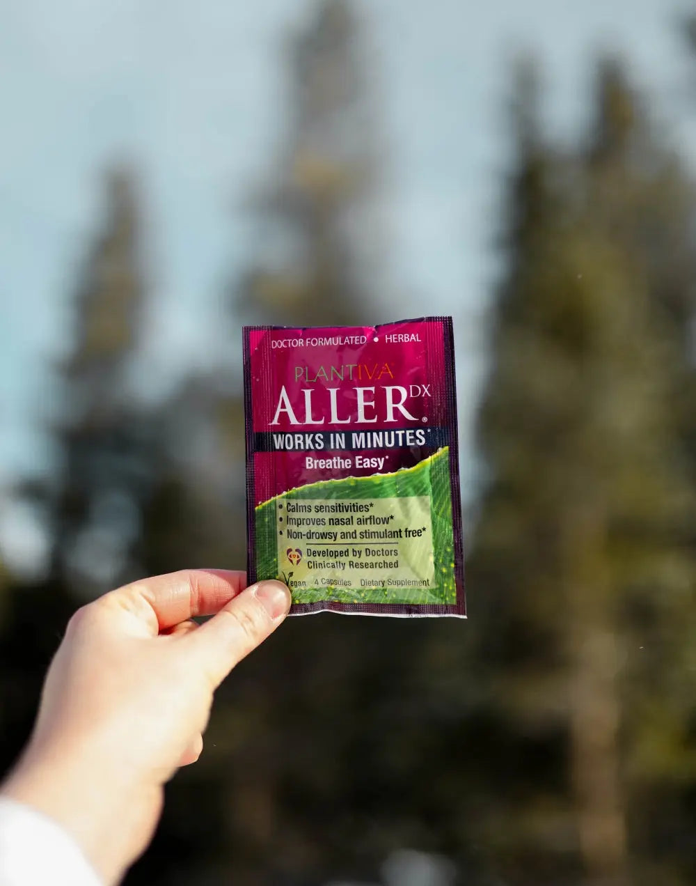 Hand holding a packet of AllerRx against a background of evergreen trees