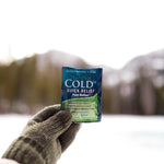 A mittened hand holding a Cold Dx packet with a snowy outdoor background