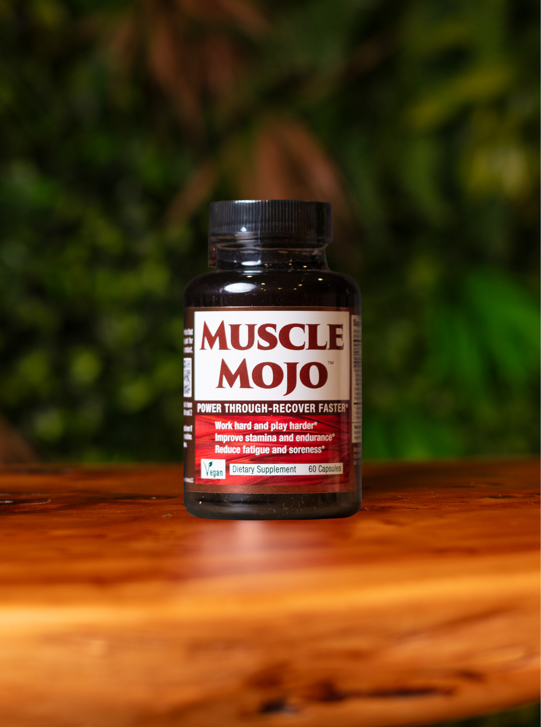 A bottle of Muscle Mojo dietary supplement placed on a wooden surface with a blurred green foliage background.
