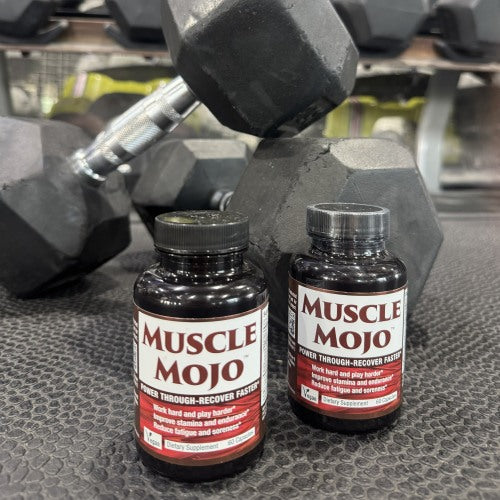 Two bottles of Muscle Mojo dietary supplement placed in front of dumbbells on a gym floor.