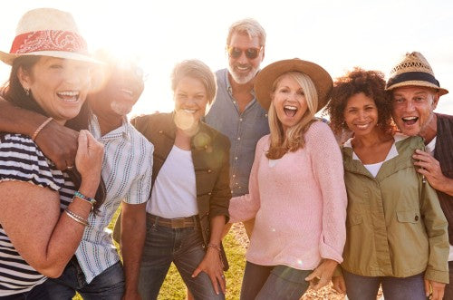 A group of seven diverse friends laughing and posing together outdoors in warm sunlight, wearing casual summer clothing and hats.